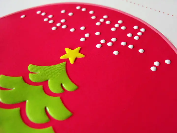 Pink sticker with festive message in braille and large type and a tactile illustration of a Christmas tree in green with a yellow star.