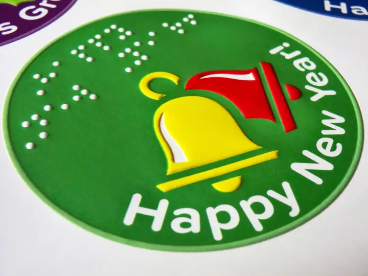 Green sticker with festive message in braille and large type and a tactile illustration of chiming bells in yellow and red.